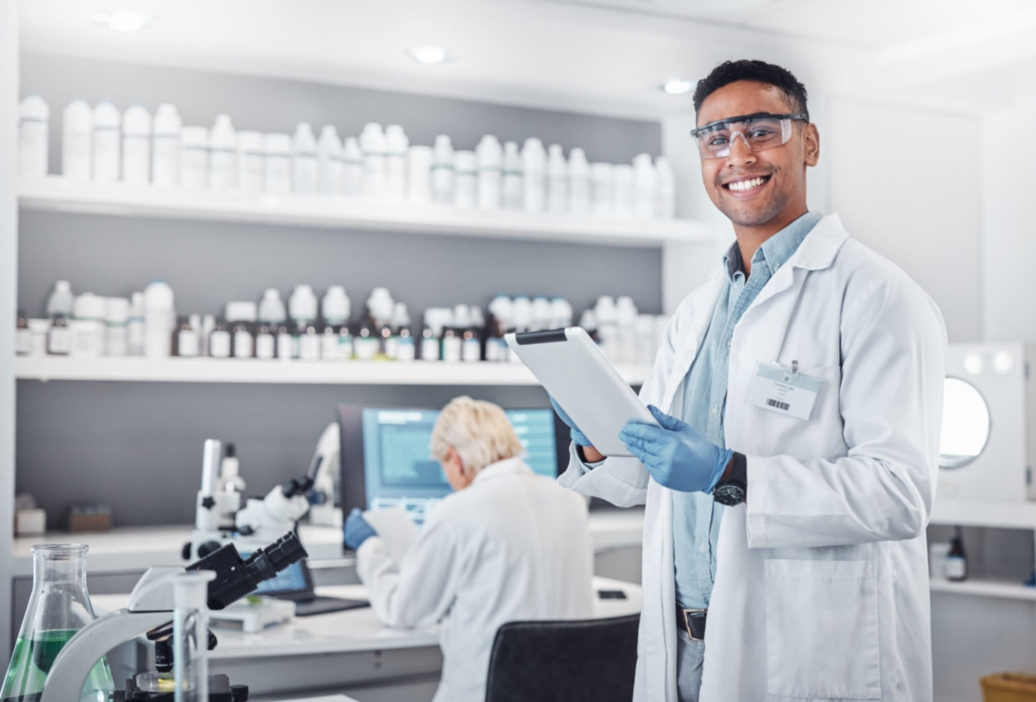 cientista sorrindo segurando sua prancheta,em um laboratório com equipamentos, atrás está mais um cientista analisando dados