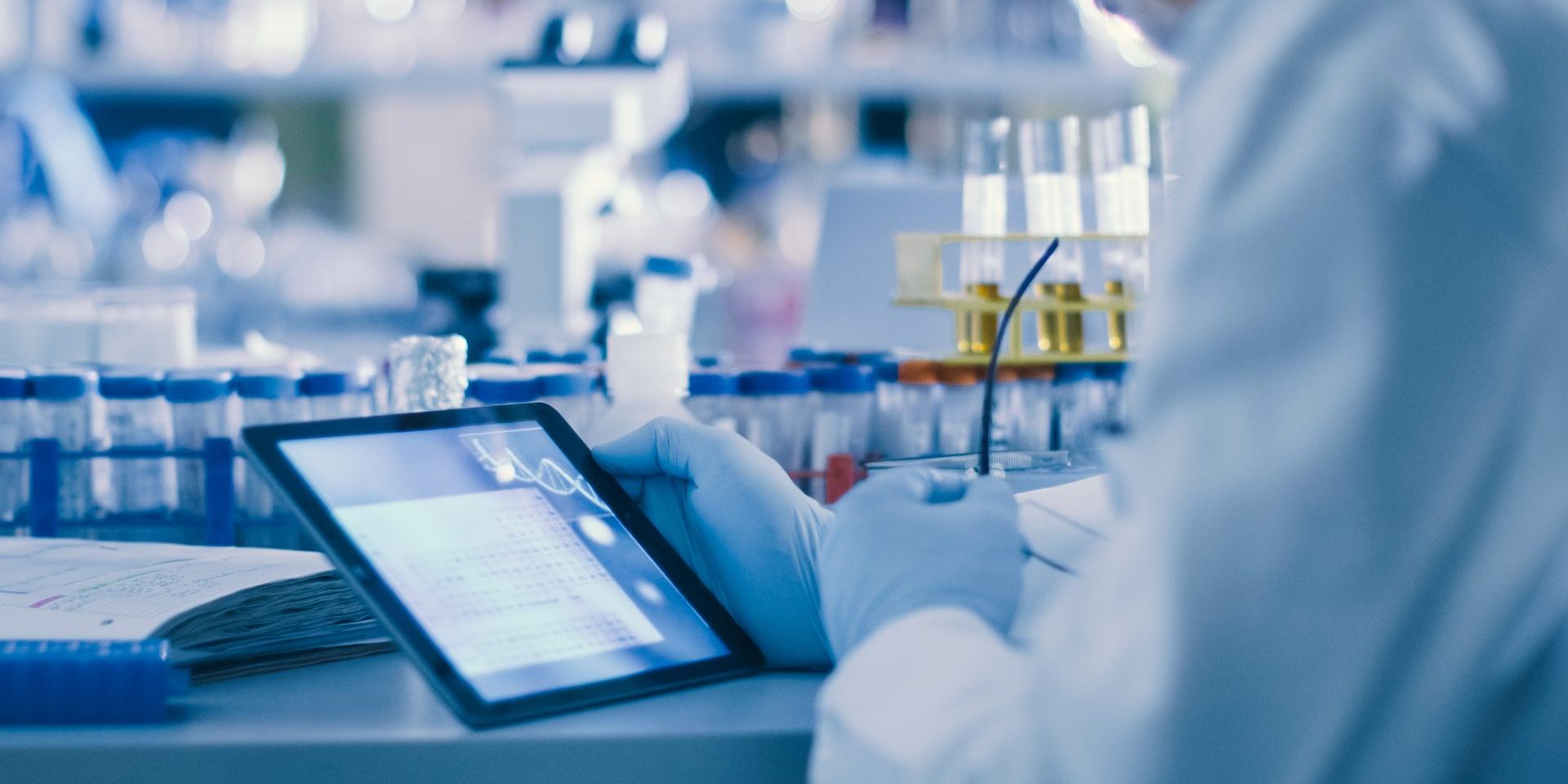 mão de cientista segura um tablet em uma mesa de laboratório, onde se encontram tubos de ensaio, microscópio e outros equipamentos laboratoriais