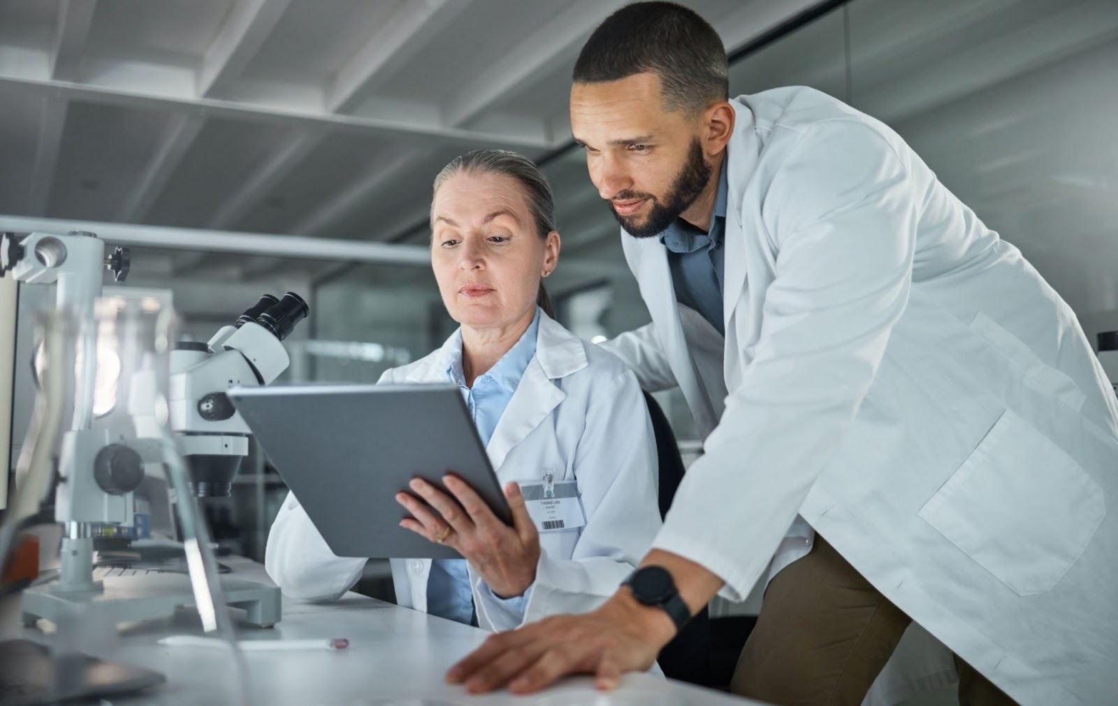 Uma cientista mulher ao lado de outro cientista homem, ambos estão olhando o tablet que está na mão da cientista, ao fundo está o laboratório