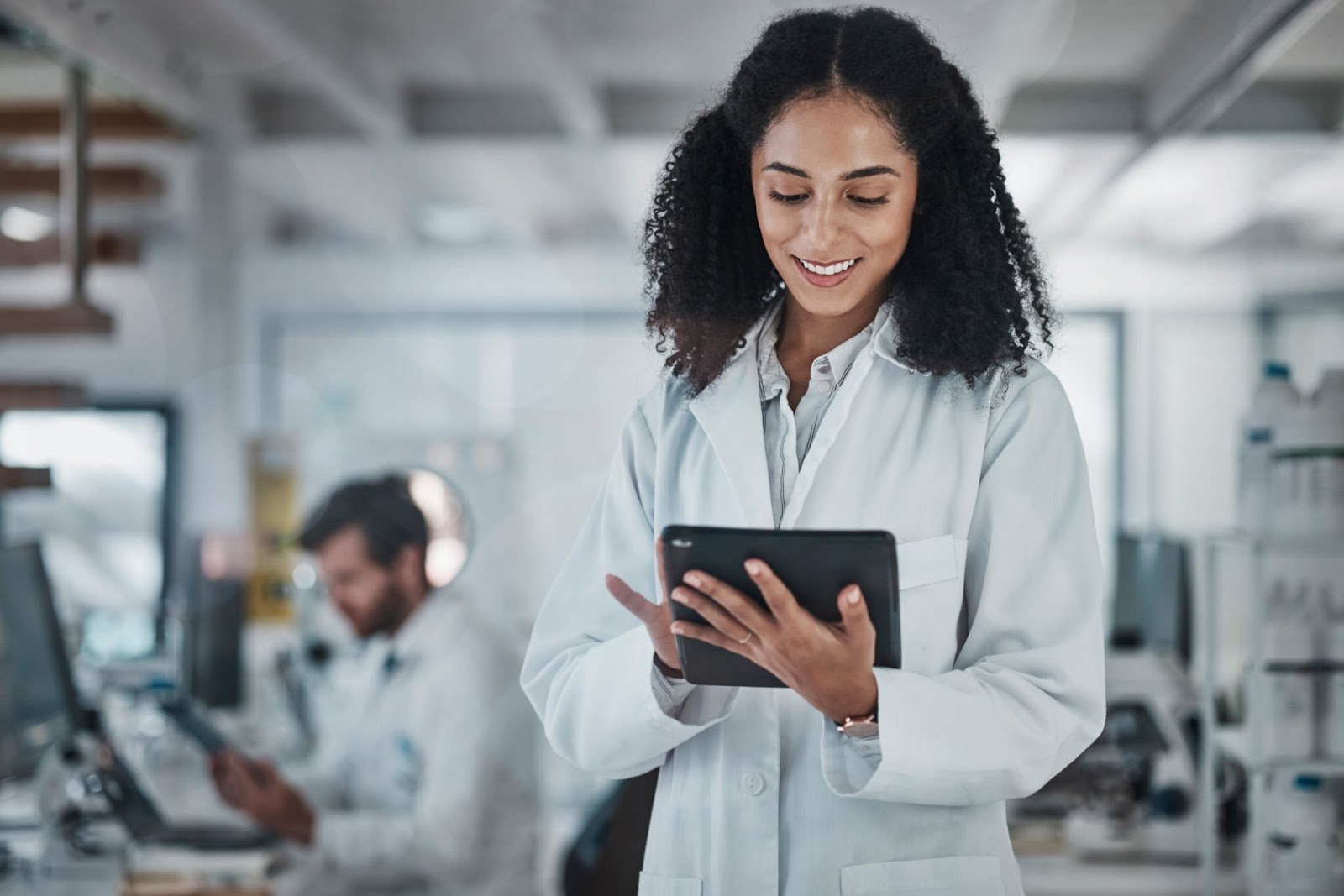 cientista negra está sorrindo e tocando em um tablet, ao fundo desfocado está um cientista homem trabalhando no laboratório